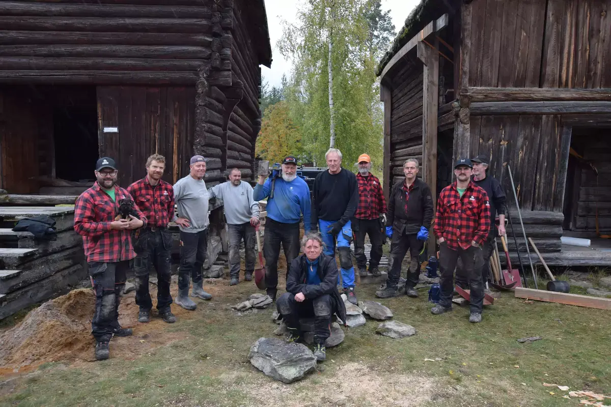 Gruppe av håndverkere samlet foran to gamle trebygninger.