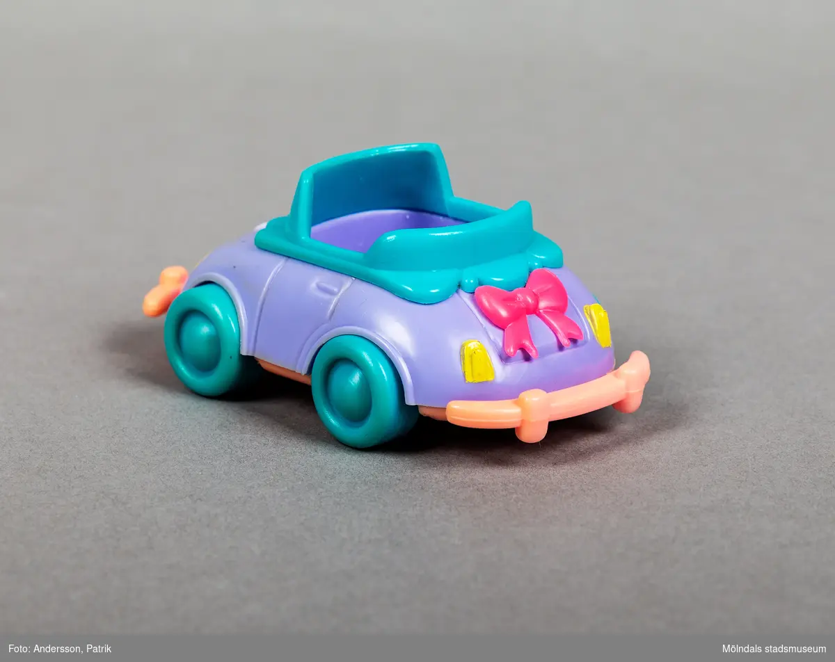 Lila leksaksbil i plast med gröna hjul och rosa kofångare. Baktill på bilen finns en rosa rosett.