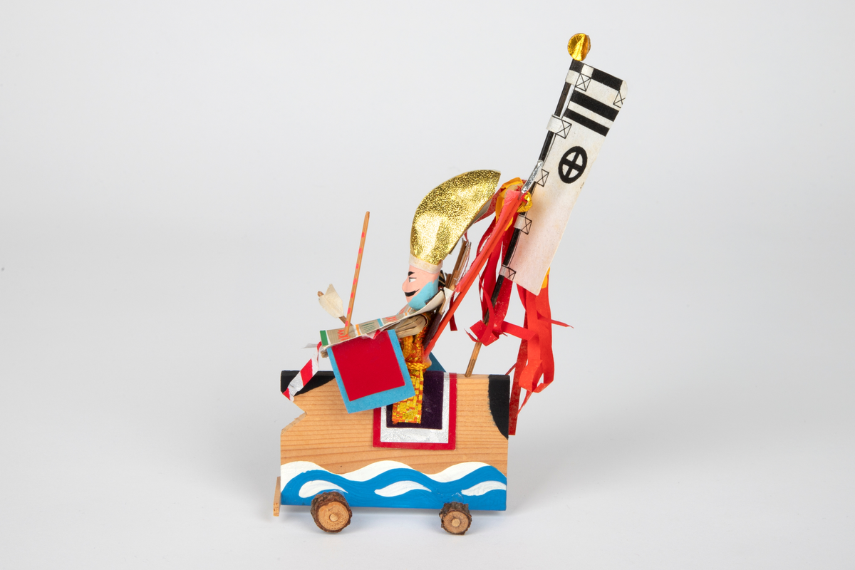 Dukke i papir festet til en vogn med hjul i tre. Dukkens hode er formet av gips. Dukken forestiller en kriger (flagbærer) med fargerik rustning og gullfarget hodeplagg. Vognen ligner en hest. Flere bannere er festet til vognen, samt piler.