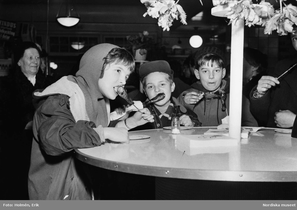Barn som äter kycklingklubbor. Demonstration av Grillman, den elektriska bordsgrillen och Findus gödkyckling på varuhuset Nordiska Kompaniet 1956.