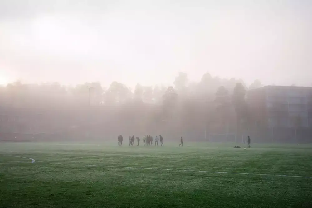 Mennesker som spiller fotball på en fotballbane i tåka. Skog og blokk i bakgrunnen.