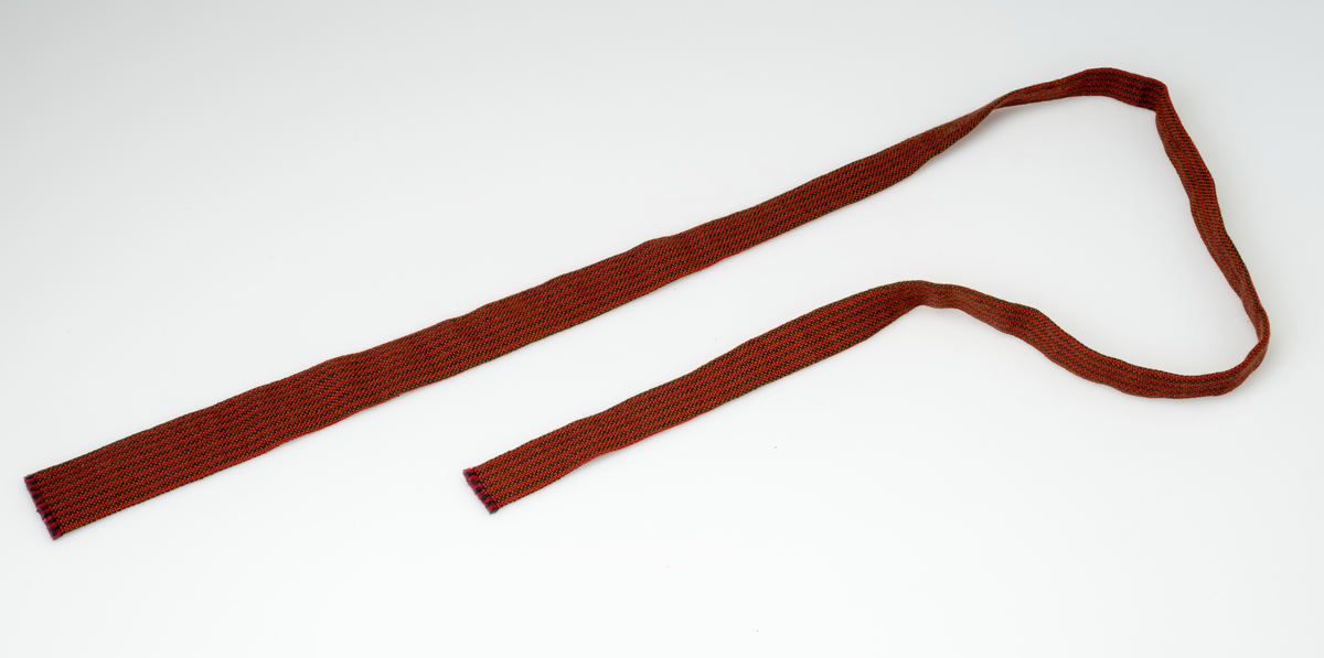 Et smalt og langt slips håndvevd av ullgarn i fargene rødt, grønt og brunt. Fargene utgjør et stripe- og rutemønster. Det vevde stoffet er brettet slik at det er dobbelt og sydd sammen med maskinsøm på vrangen. I hver ende av slipset er det korte frynser (0,2-0,3cm). Slipset er smalere i den ene enden enn den andre.