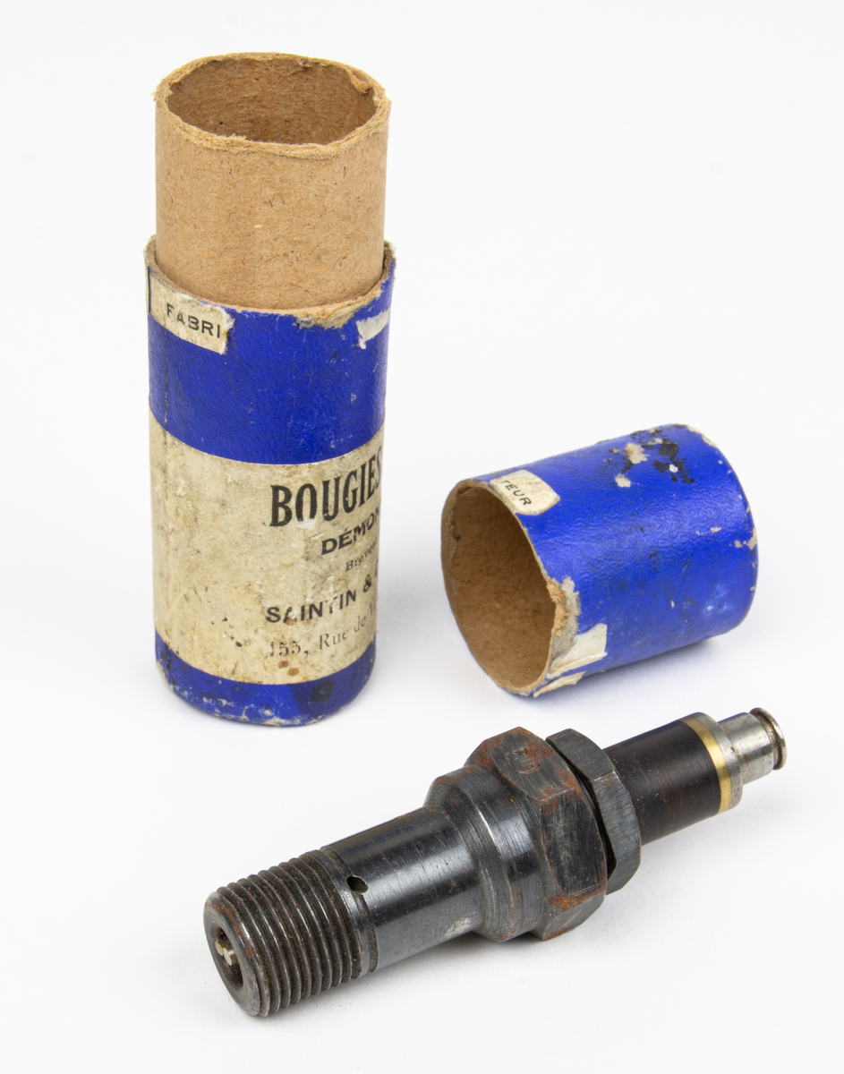 Tändstift, av fabriketet Saintin. Förvaras i originalförpackning i form av ett blått papprör med etikett med bland annat texten Bougies Saintin och dateringen 1927. I förpackningen följer en dokument. (Tändstift för motorer med hög kompression och hög hastighet.)