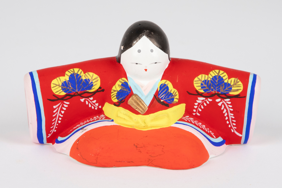 Sittende prinsessefigur i gips med utstrakte hender. Figuren er kledd i en rød og oransje kimono med mønster i blå, gul, gull, hvit og svart og gult belte. Dukken har en svart lang flette bak.