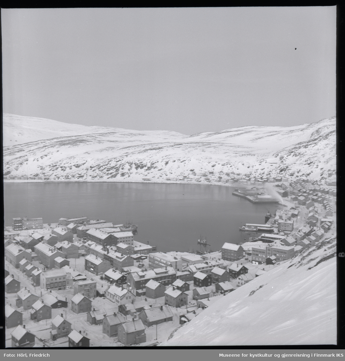 Utsikt over Hammerfest sentrum fra Salenfjellet i vinteren 1957/1958. Mesteparten av byen er gjenreist og man ser mange sentrale bygg som kommunehuset, Handelshuset Nissen, rådhuset, bankbygget, Findusfabrikken og Hauans materialhandel. I bakgrunnen ligger bydelen Molla og det snødekkede fjellandskapet.