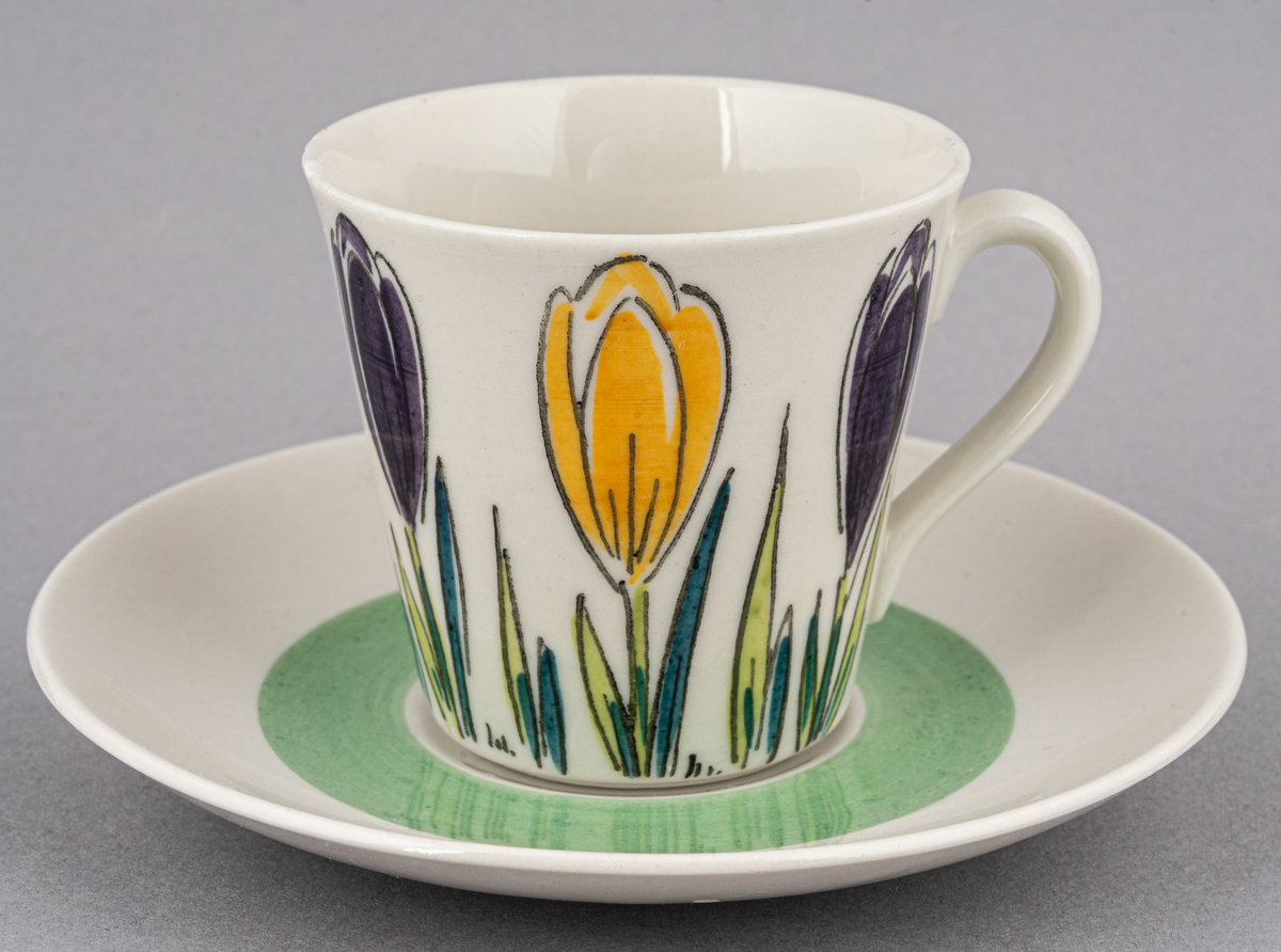 Kaffegods i flintgods med rikt handmålad dekor i form av krokusar i gult och lila, modell E. Dekoren skapad av Ellen Mortensen 1959, tillverkades till 1964. Koppen har två nagg vid kanten.