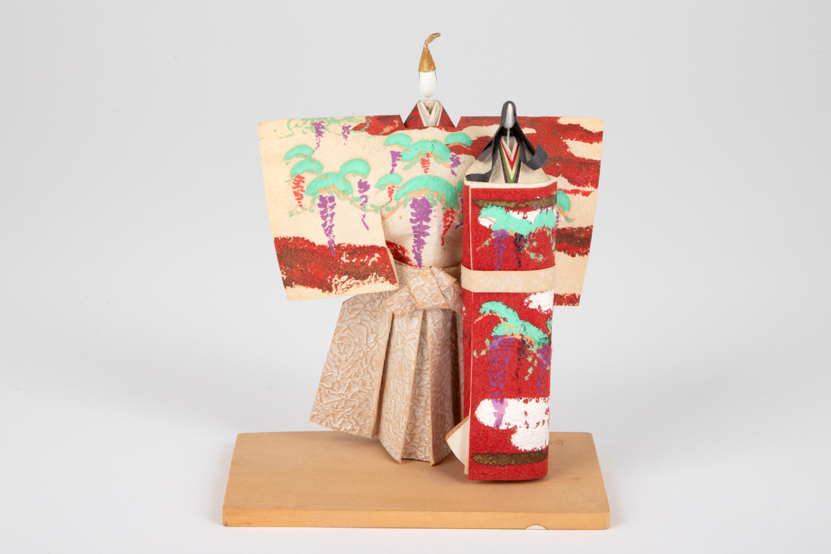 Dukkepar (kvinne og mann) i papir montert på en treplate. Dukkene er kledd i kimonoer i sølv, rustrød og beige med mønster i lilla, hvit, oker og turkis.