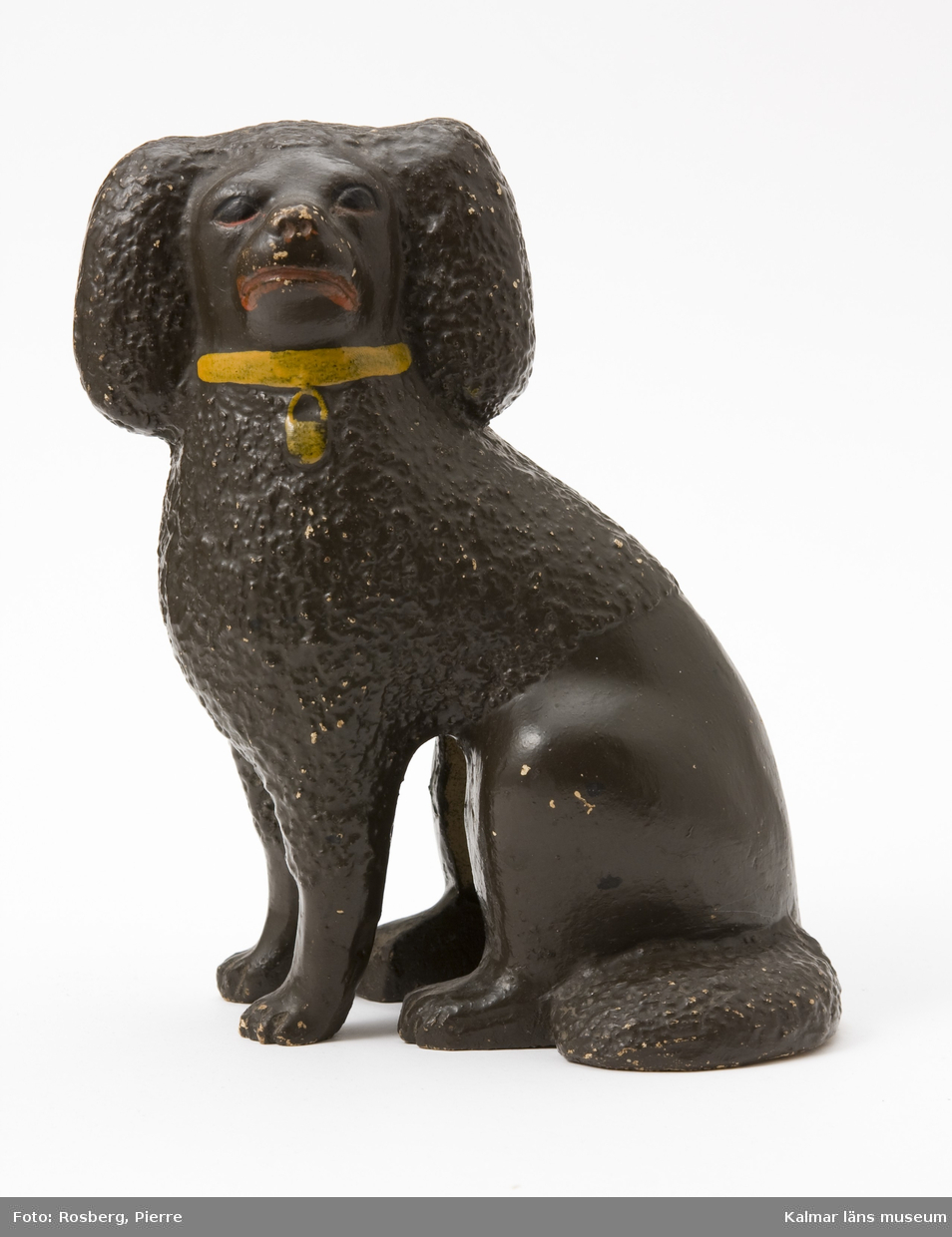 KLM 12119. Figurin. Tillverkad av lergods. Figurin i form av en sittande hund, pudel, brunfärgad, med gult halsband, röd mun och nos, ögon i svart och rött. Stämpel: Augusta, JB.  Par med KLM 12118.
