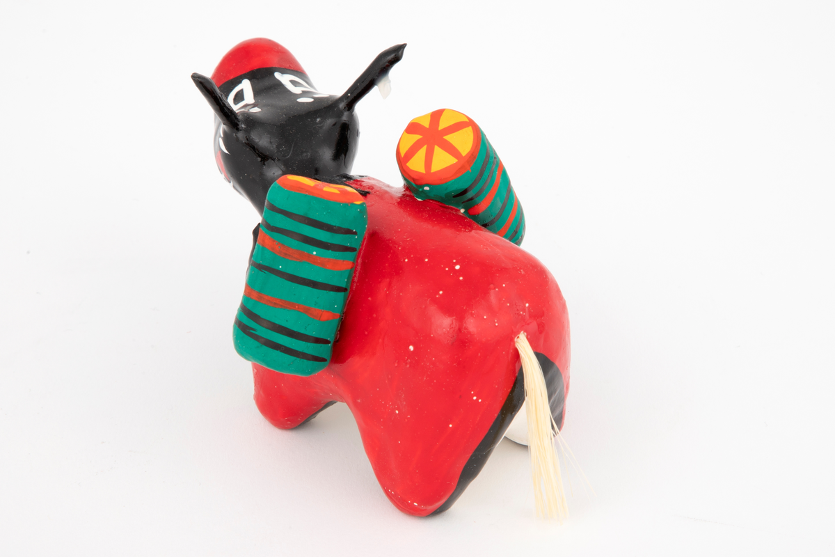 Okse/vannbøffel i pappmasjé med dinglehode. Figuren er rød med svart, rødt og hvitt hode. Den bærer to vesker i grønt, svart, oransje og gul på ryggen.
