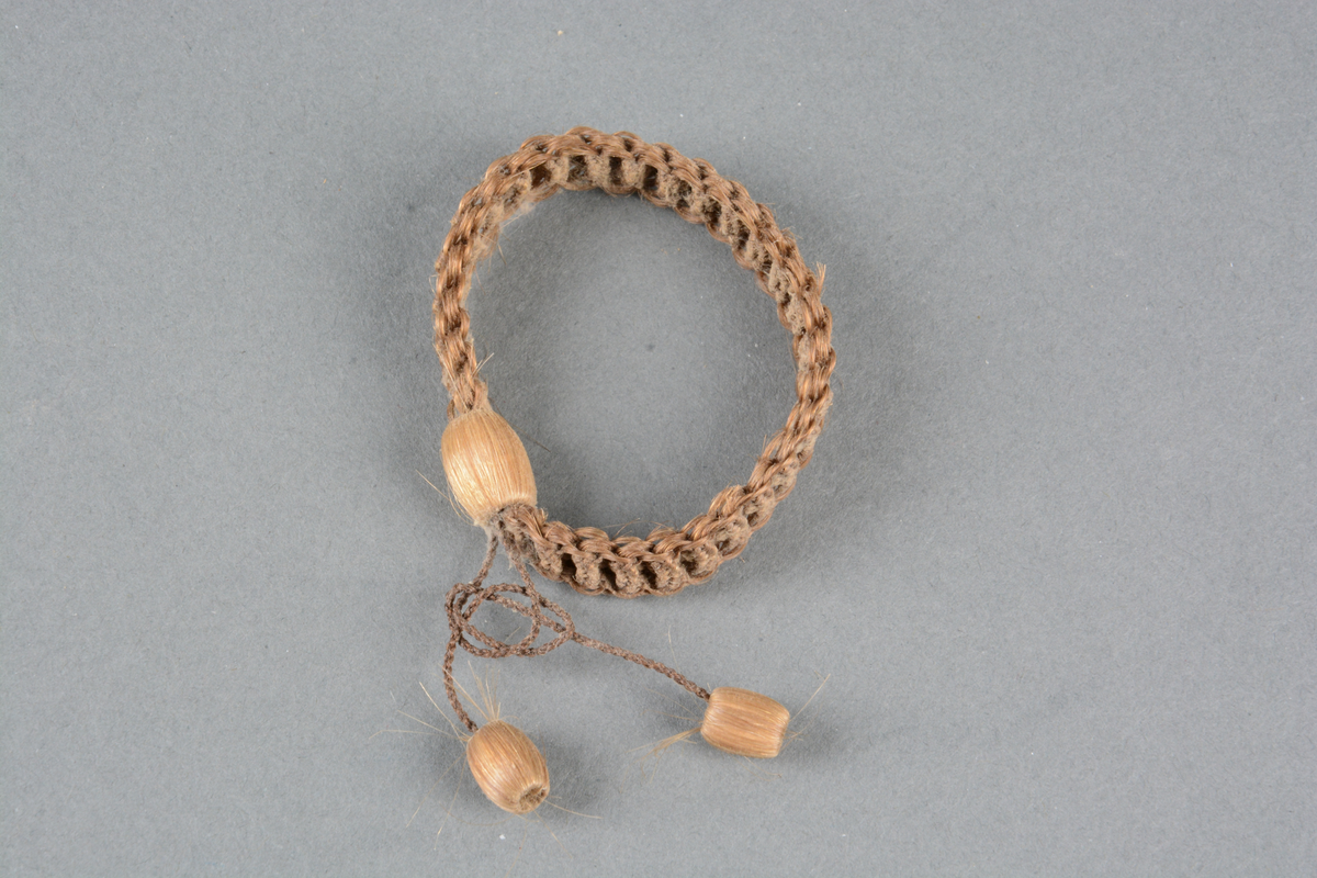 Armbånd av hår, til barn (?). Vaffelflettet bånd, samlet i hårtrukket perle. To mindre perler henger ved.