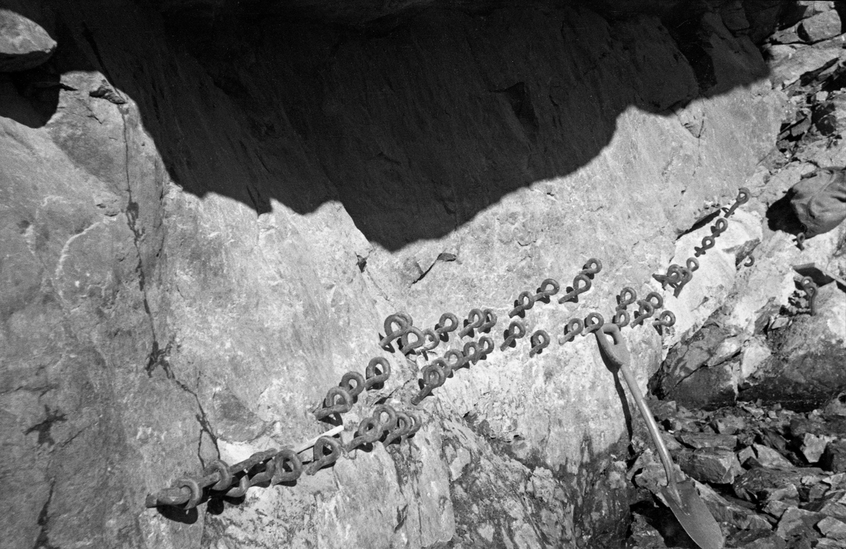 Fra bygginga av Sølvstudammen i Tune i Østfold i 1936. Fotografiet er tatt fra et stillas ned mot bergflata ved elveleiets ytterkant. Her ser vi hvordan anleggsarbeiderne har drevet inn en masse bolter med oppstikkende «øyne», antakelig et forankringstiltak for det som etter hvert skulle bli dammens styrtgolv. Under boltrekkene står det en spade mot bergveggen. Sølvstudammen ligger i Ågårdselva, et cirka fem og en halv kilometer langt vassdrag som renner fra den nordvestre enden av Isnesfjorden (Vestvannet) i Nedre Glommas vestre løp, sørvestover gjennem berglendt terreng mot innsjøen Visterflo. Høydeforskjellen mellom Isnesfjorden og Visterflo er på bortimot 25 meter. Ågårdselva har tre fossefall, det øverste her ved Sølvstu, deretter ved Valbrekke og nederst ved Solli. Behovet for den dambygginga vi ser på dette fotografiet var forårsaket av selskapene Borregaards og Hafslunds inngrep i Glommas østre løp, hovedløpet, som gjorde det svært vanskelig å få tømmer som skulle til bedrifter nedenfor Sarpsfossen via Mingevannet, Isnesfjorden og tømmertunnelen til Eidet gjennom det trange sundet ved Trøsken (mellom Mingevannet og Isnesfjorden) i lavvannsperioder. Dette forsøkte man å løse ved å heve vannspeilet i Isnesfjorden med en dam som skulle plasseres 10-15 meter ovenfor den eksisterende dammen ved Sølvstufossen. Se mer informasjon under fanen «Opplysninger».