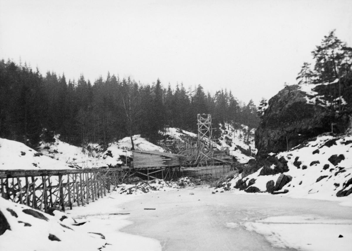Fra bygginga av en ny dam ved Sølvstufossen, øverst i Ågårdselva i Tune i Østfold, som er en del av Nedre Glommas vestre løp. Anleggsarbeidet startet vinteren 1936, i en periode da det knapt rant vann i elveløpet. Dette fotografiet er tatt i motstrøms retning. Rampa med skinnegang langs nordre elvebredd (til venstre i bildet) ble brukt til å trille materialer og redskap i vogner til og fra anleggsområdet. Inntil denne rampa hadde anleggsarbeiderne reist et bordskur med pulttak. Bakenfor skimter vi forskalinger til dammurer, og over det hele rager noe som må antas å ha vært et heistårn. Ågårdselva er et cirka fem og en halv kilometer langt vassdrag som renner fra den nordvestre enden av Isnesfjorden (Vestvannet) i Nedre Glommas vestre løp, sørvestover gjennem berglendt terreng mot innsjøen Visterflo. Høydeforskjellen mellom Isnesfjorden og Visterflo er på bortimot 25 meter. Ågårdselva har tre fossefall, det øverste her ved Sølvstu, deretter ved Valbrekke og nederst ved Solli. Behovet for den dambygginga vi ser på dette fotografiet var forårsaket av selskapene Borregaards og Hafslunds inngrep i Glommas østre løp, hovedløpet, som i lavvannsperioder gjorde det svært vanskelig å få tømmer som skulle til bedrifter nedenfor Sarpsfossen via Mingevannet, Isnesfjorden og tømmertunnelen til Eidet gjennom det trange sundet ved Trøsken. Dette forsøkte man å løse ved å heve vannspeilet i Isnesfjorden med en dam som skulle plasseres 10-15 meter ovenfor den eksisterende dammen ved Sølvstufossen. Se mer informasjon under fanen «Opplysninger».