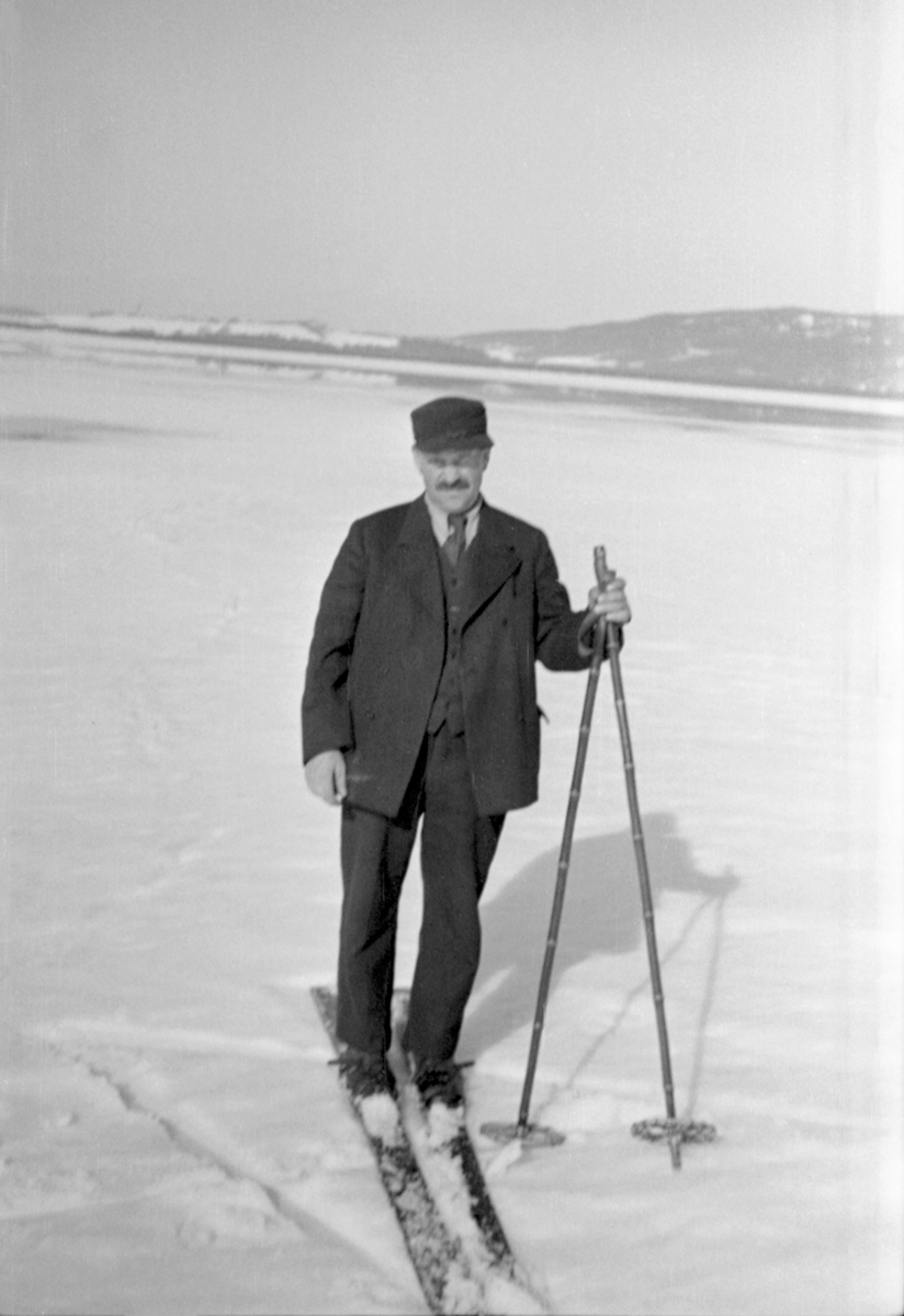 Fløtingsformann Carl Sand (1879-1952) fra Enebakk, fotografert på ski ved Årnestangen, på vestsida av innsjøen Øyerens nordende, i Rælingen. Sand var kledd i dress med vest og slips, og han hadde ei skyggelue på hodet.
