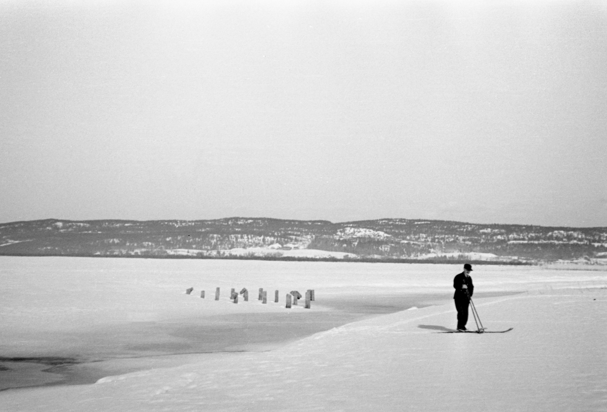 Fløtingsformann Carl Sand (1879-1952) fra Enebakk, fotografert på ski ved Årnestangen, på vestsida av innsjøen Øyerens nordende, i Rælingen. Sand var kledd i dress med vest og slips, og han hadde ei skyggelue på hodet. Bak Sand, sentralt i bildet, stakk det opp en del pælestubber, antakelig etter en fløtingsinnretning.