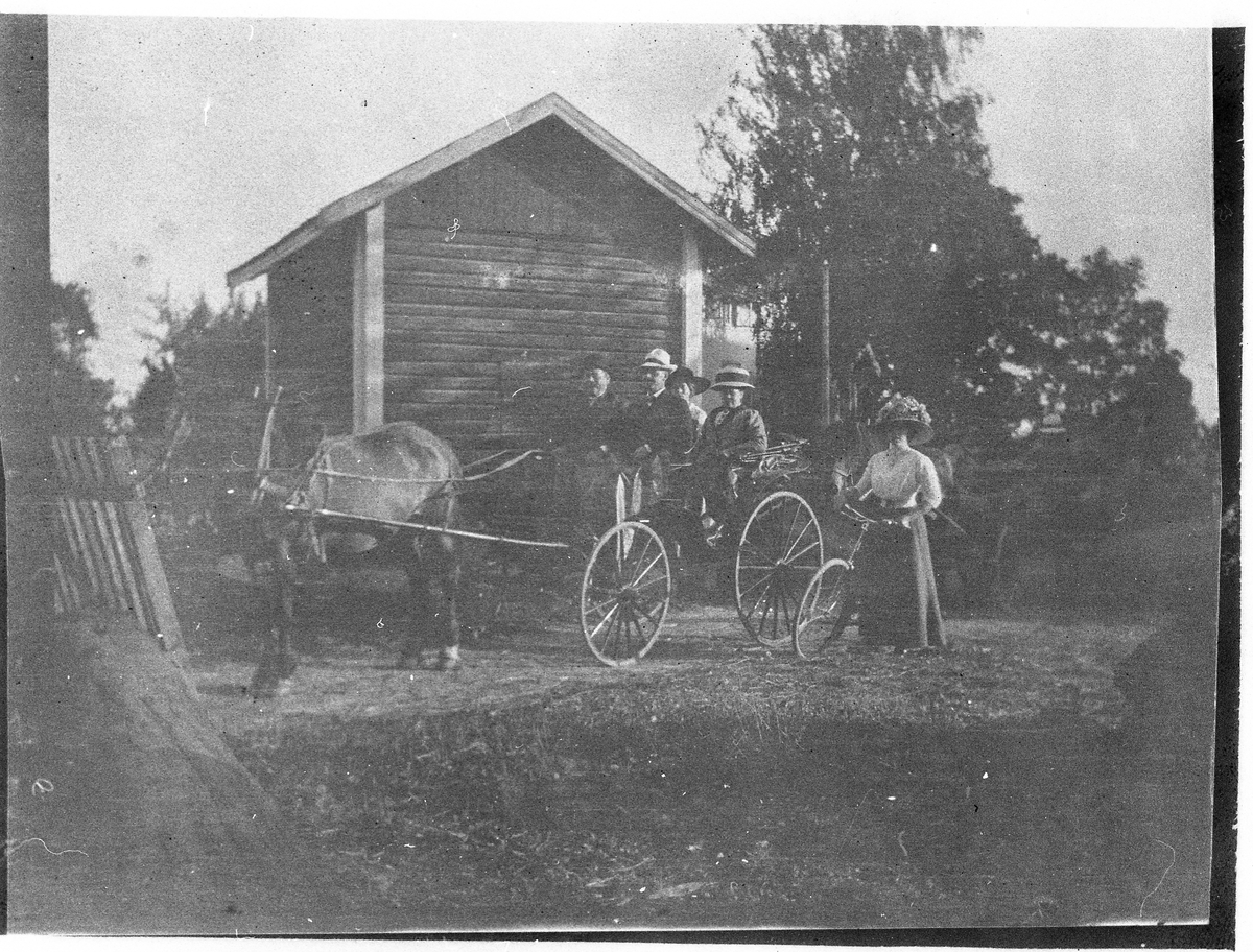 En syklist og ei hestekjerre med personer foran en mindre tømmerbygning. Ingen informasjon foreligger.