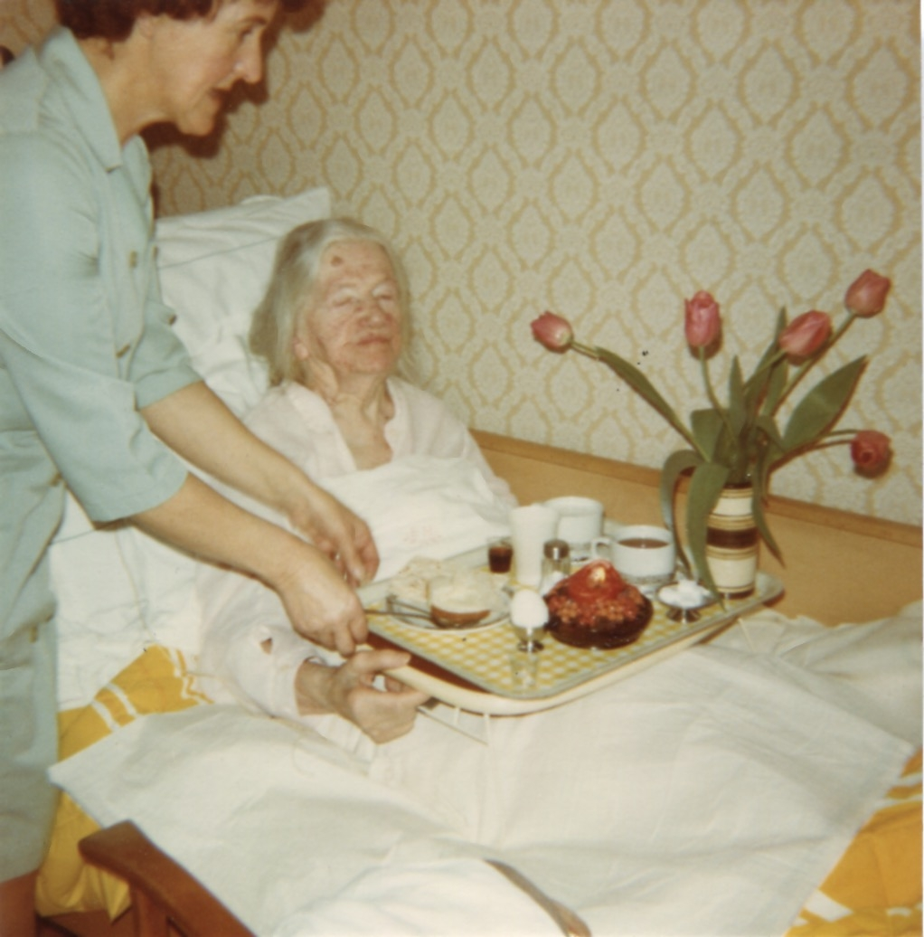 Vårdbiträde Maj Britt Reimertz ställer fram en bricka till Wida Ahlström (1891 - 1981) som är sängliggande. Troligtvis en uppvaktning då det finns frukost, ljus och tulpaner på brickan.