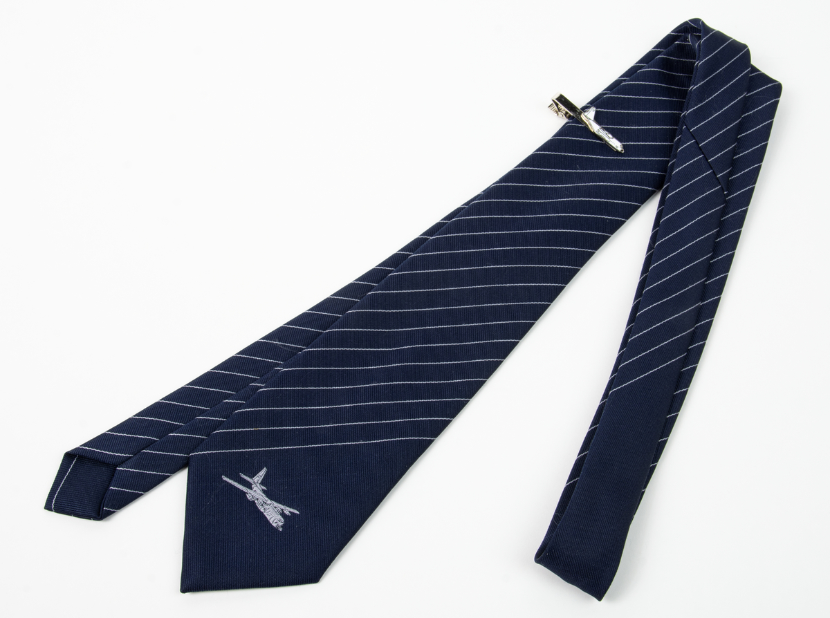 Blå slips med smala vita diagonala ränder och en bild på flygplan Tp 84. Till slipsen hör en slipsnål även där med en bild på flygplan Tp 84.
