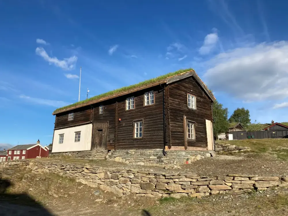 Bildet viser et bygg i laftet tømmer og mur. I forgrunnen er det ens steinmur. I bakgrunnen er det blå himmel og andre hus skimtes.