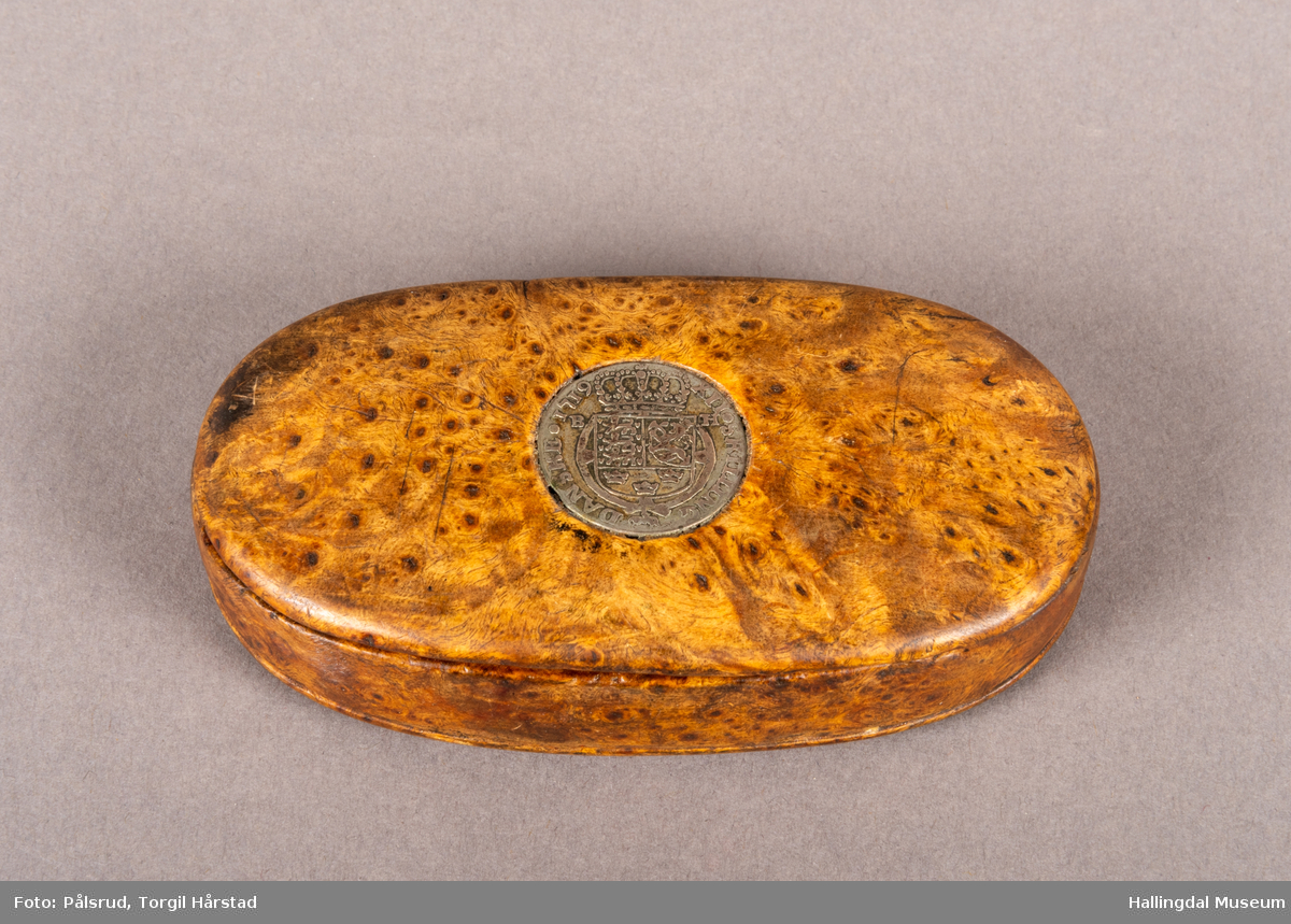 En brun, oval snusdåse i valbjørk. Lokket er festet med trehengsler på den ene langsiden. Innfelt i lokket sitter en 12-skilling fra 1719. Under bunnen er det innskåret "K O S ANO 1642".