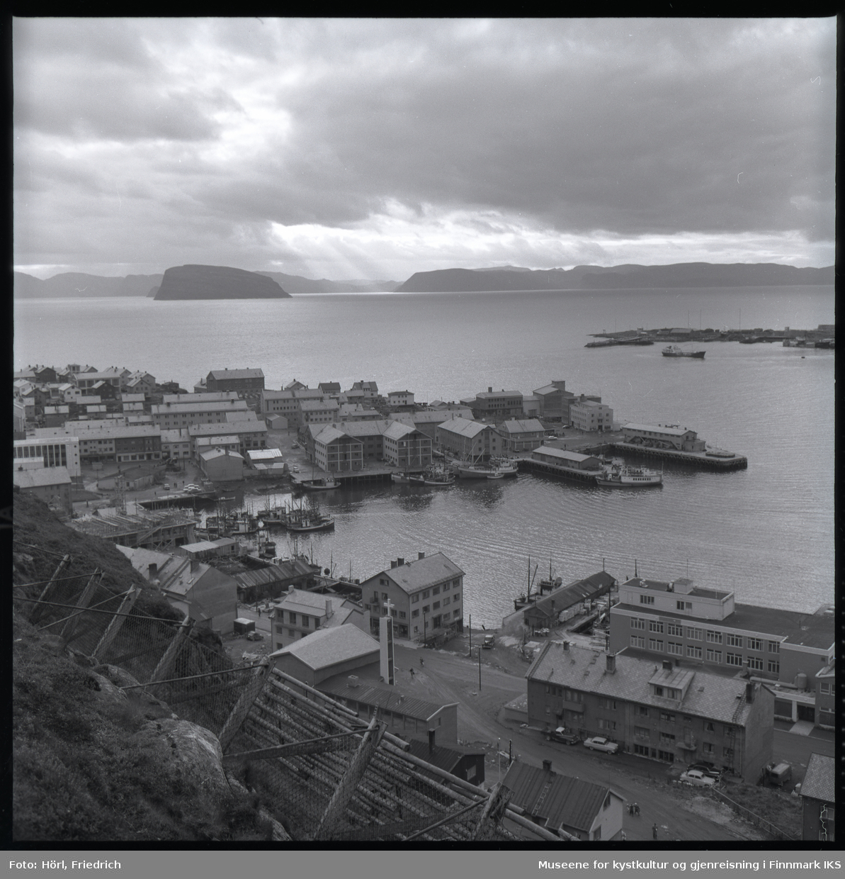 Utsikt over Hammerfest sentrum og havn fra Salenfjellet. I havna ligger ulike type båter. Man ser gjenreiste bygg, deriblant den katolske kirka St. Mikael, men også brakker og bygninger som er under oppføring. I fjellsiden i forgrunnen ser man gjerder til rassikring. Til høyre i bilde ser man spissen til Fuglenes. I bakgrunnen ser man Sørøysundet med øyene Håja og Hjelmen, samt silhuetten av Sørøya.