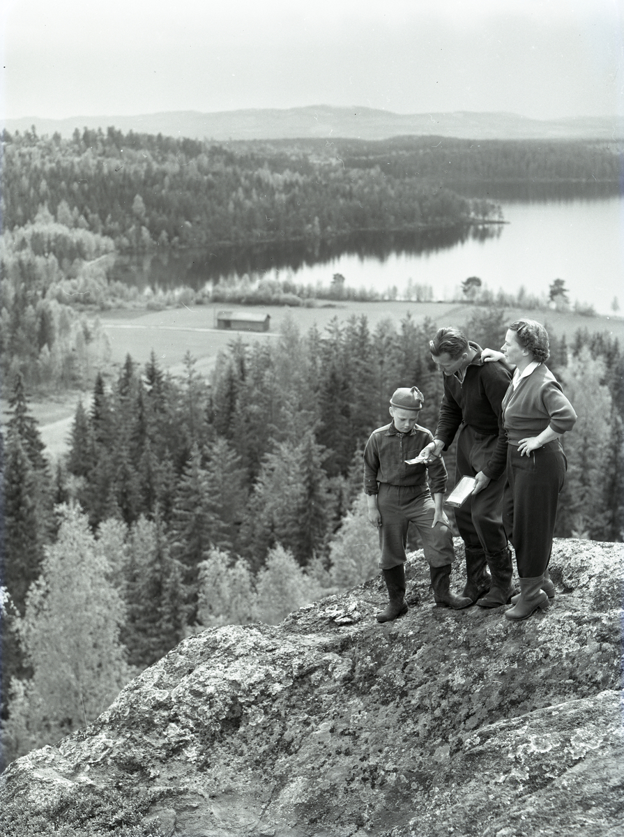Hilding Mickelsson står på ett berg med sin fru och son hållandes karta och kompass i händerna. Bakom den orienterande familjen syns en landskapsvy med lada, skog och vattendrag.