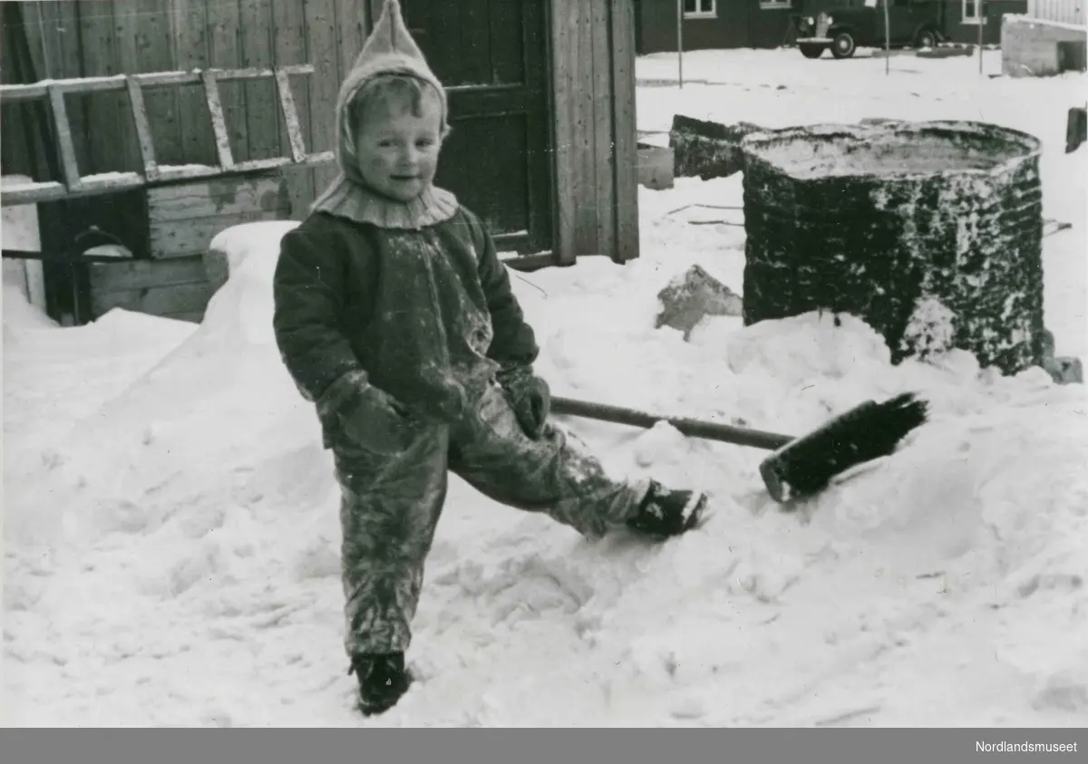 Et lite barn med strikket lue står ute i snøen. Bildetekst: Bakgård i Dronningens gate 3".