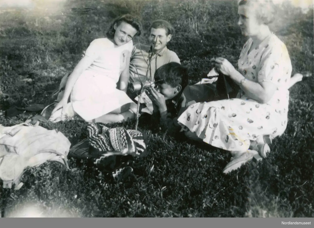 Fire mennesker sitter ute i lyngen. En ung kvinne og en ung mann smiler mot fotografen. En gutt ser i en kikkert. En voksen dame sitter og strikker. På bakken foran dem ligger en strikkejakke. Bildetekst: Fra det Nygårdsjønske fangenskap 1942".