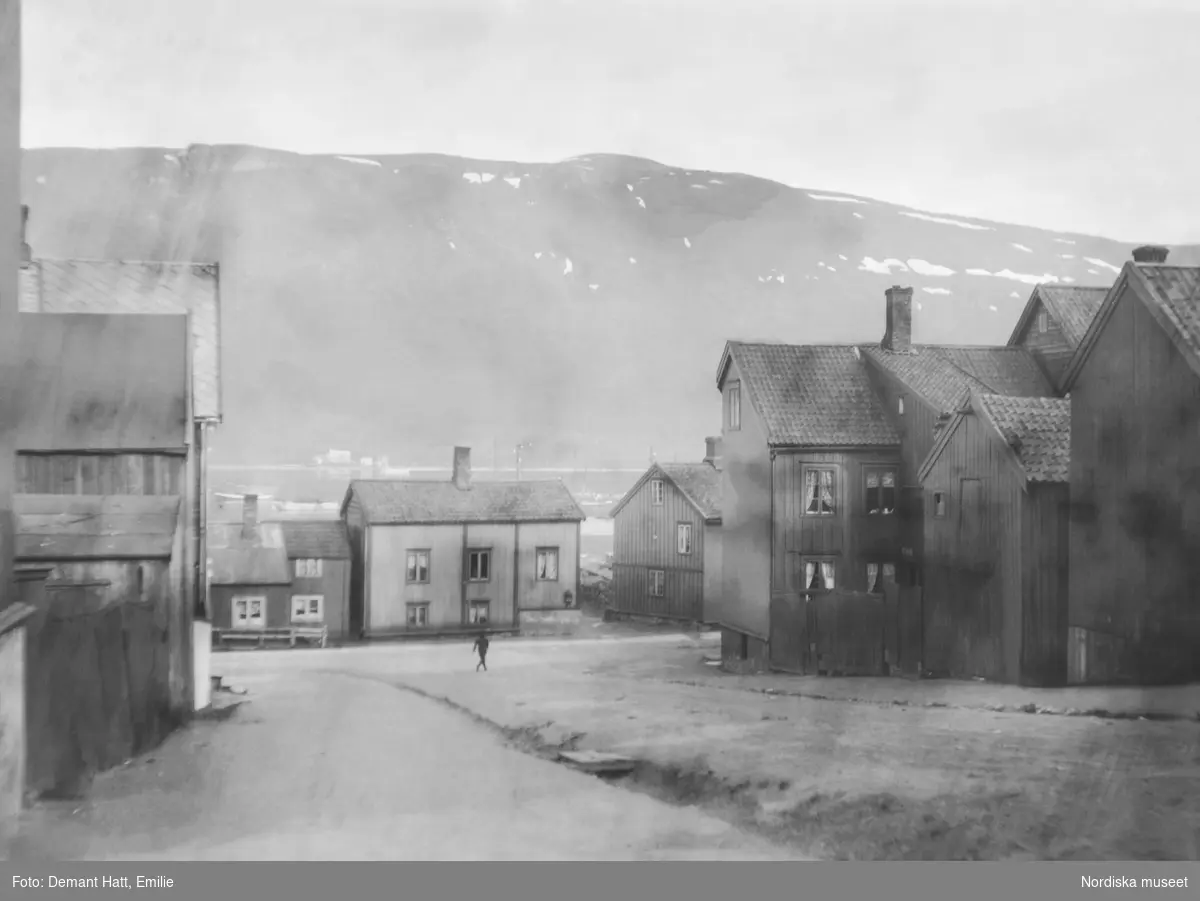Stadsbild från Tromsø i Norge. Låga trähus kantar gatorna, i bakgrunden anas ett fjäll. En ensam människa står på en gräsbevuxen yta. Bilden ingår i en serie fotografier tagna av Emilie Demant Hatt i Sapmi mellan åren 1907 och 1916.