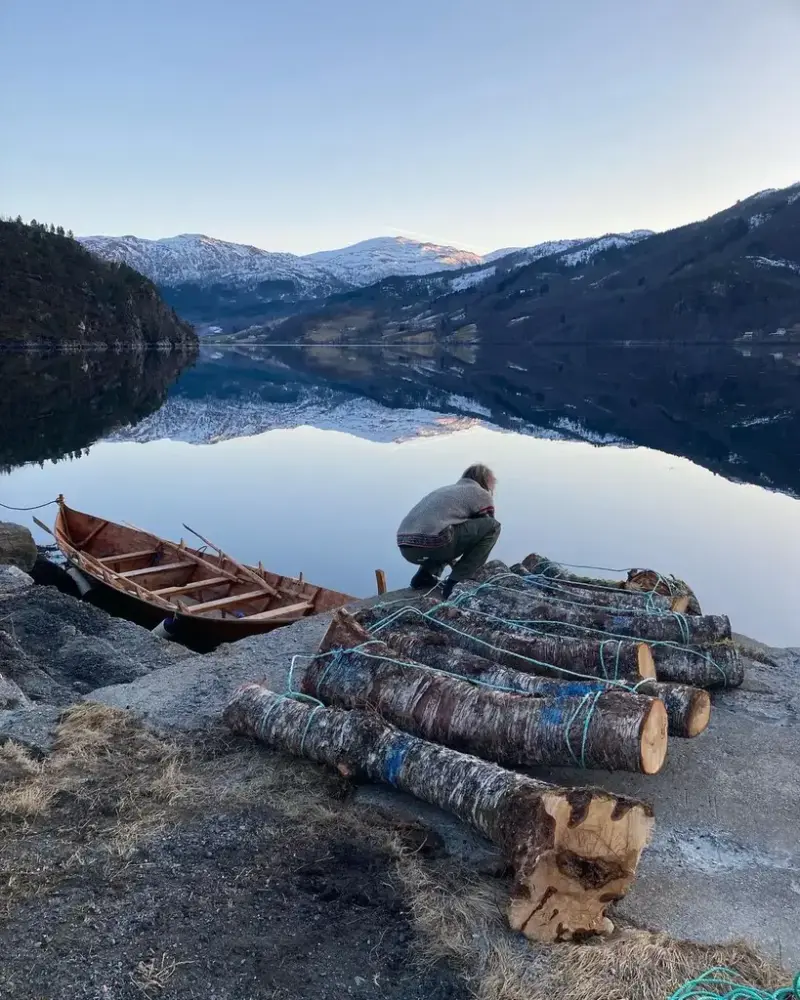 En av museets håndverkere står på kaia med trestokker. Ved siden av en robåt..