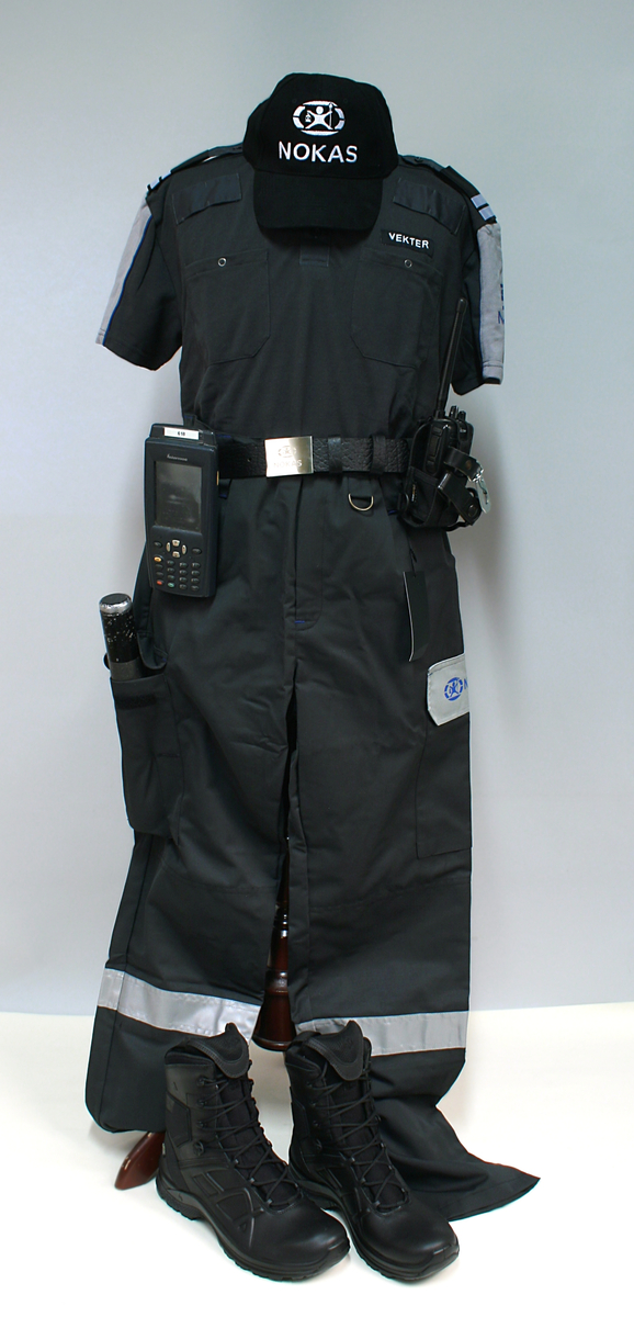Sort uniform bestående av kortermet poloskjorte, bukser og skyggelue med Nokas-logo, og tilhørende belte rigget med lommelykt og walkie-talkie.