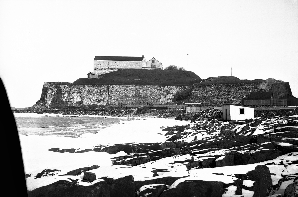 Vykort, "Varberg - Fästningen." Vinterbild av fästningen sedd söderifrån. På stranden ses stenhuggarbaracker som hör till stenbrottet nedanför fästningen.
Bild 1: sv/v högre upplösning, plåten trasig i vänster kant. Bild 2: vykort sepiaton.