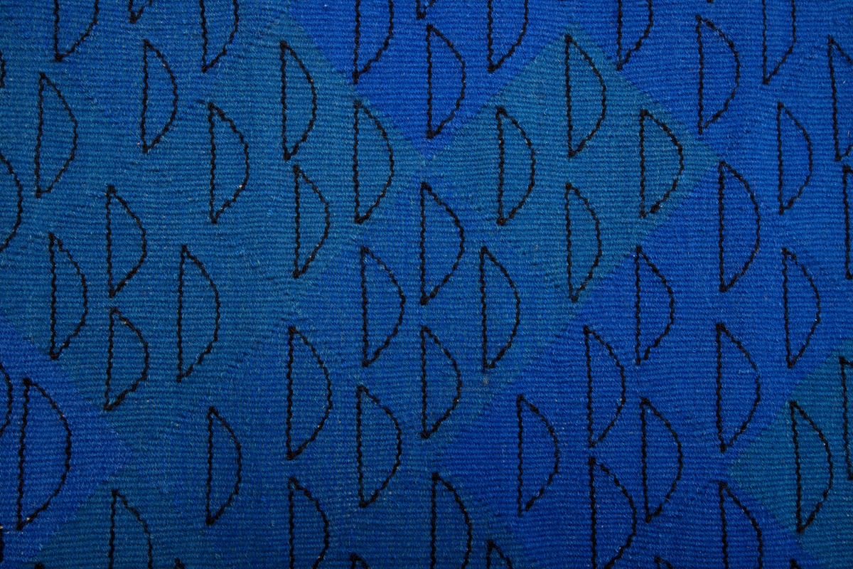 Håndvevd rektangulært billedvev i ull med geometrisk motiv i svart, blått, oransje og beige. Langs venstre og høyre kant er renningstrådene flettet og festet på baksiden med påsydde vevde bånd.
