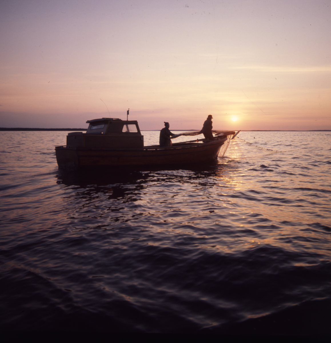 Två personer i en fiskebåt ute på vattnet. Solen står lågt på himlen.