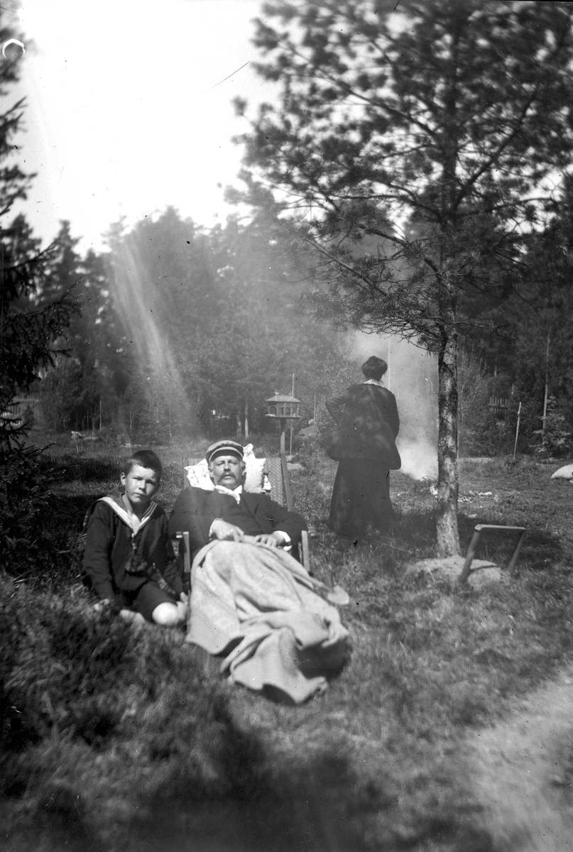 En familj i skogen. Mannen ligger i en vilstol med en filt över benen. En pojke sitter på marken bredvid honom och en kvinna syns i bakgrunden vänd mot en eld..