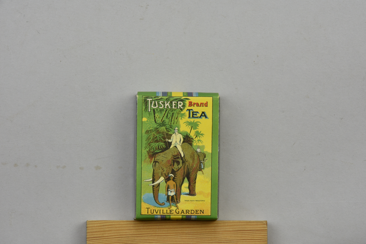 Teförpackning med motiv av två män i tropikhattar på elefanter samt en elefantförare. märkt "Tusker brand tea, Tuville Garden" samt "Trade mark registered". Framsida.