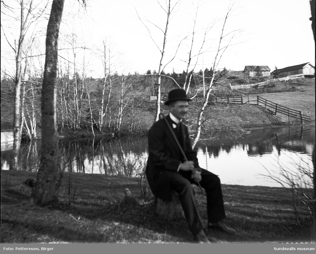 En man med hatt, käpp och helgdagskläder sitter på en stubbe i lantlig omgivning. Bakom syns ett stilla vattendrag, inhägnader i trä och enklare byggnader, kanske en fäbodvall.
