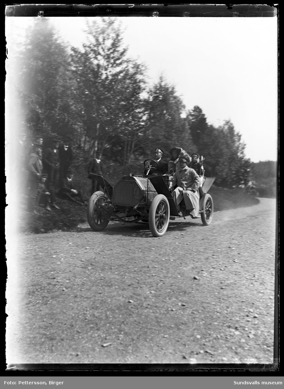 En ålderdomlig öppen bil utan vindruta på en landsväg, troligen i Njurundatrakten. I bilen sitter fyra personer och vid vägkanten står en grupp män och beundrar fordonet. Bilden är tagen vid en biltävling som Svenska Motorklubben (SMK) anordnade sommaren 1909 på sträckan Haparanda-Stockholm. Bilen är en 40 hästkrafters Horch från 1907 som ägdes av firman Bil-Bolaget.