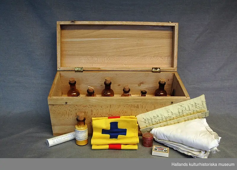 Sjukvårdslåda i trä från tidigt 1900-tal. Lådan innehåller två armbindlar, två mitellor, en leukoplastrulle, kloramintabletter, ask för säkerhetsnålar "The Globe", en glasflaska med natriumbikarbonat, 