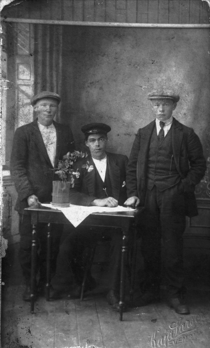 Studioportrett av tre menn, en sitter ved et bord og de to andre står ved siden.