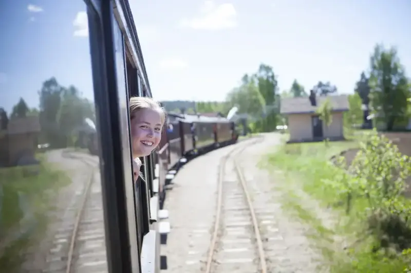 Jente kikker ut av togvinduet i en av Tertittens passasjervogner.  I bakgrunnen skimtes damplokomotivet og stasjonsbygningen på Fossum stasjon.