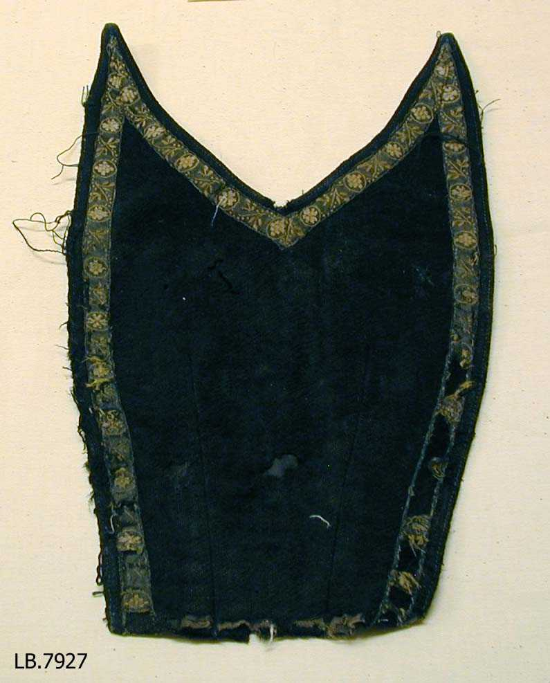 Bringeklut i svart/mørkeblå (?) ulltøy pynta med mønstra band i bomull. Hekter (rusta) på innsida venste side.