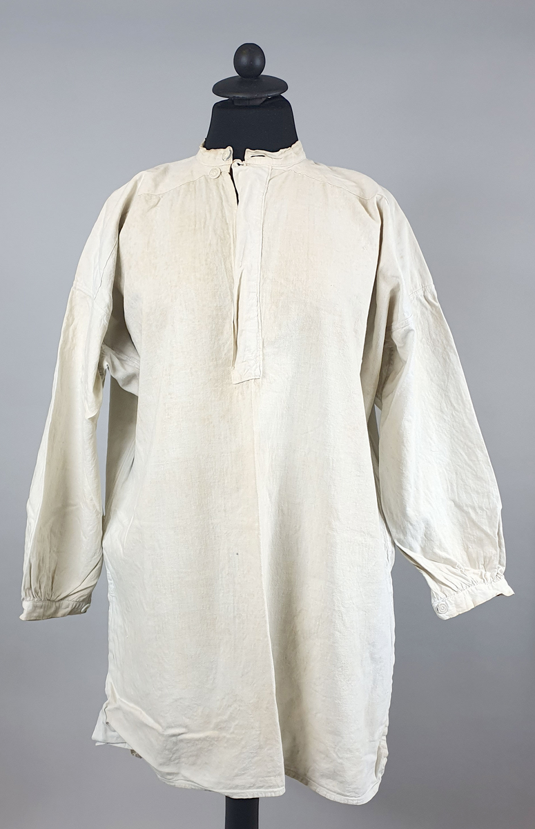 Skjorte (busserull) av bomull, med åpning foran og påsydd knappestolpe med knapp øverst. Stående krage med knapp. Smale mansjetter med kanpp.