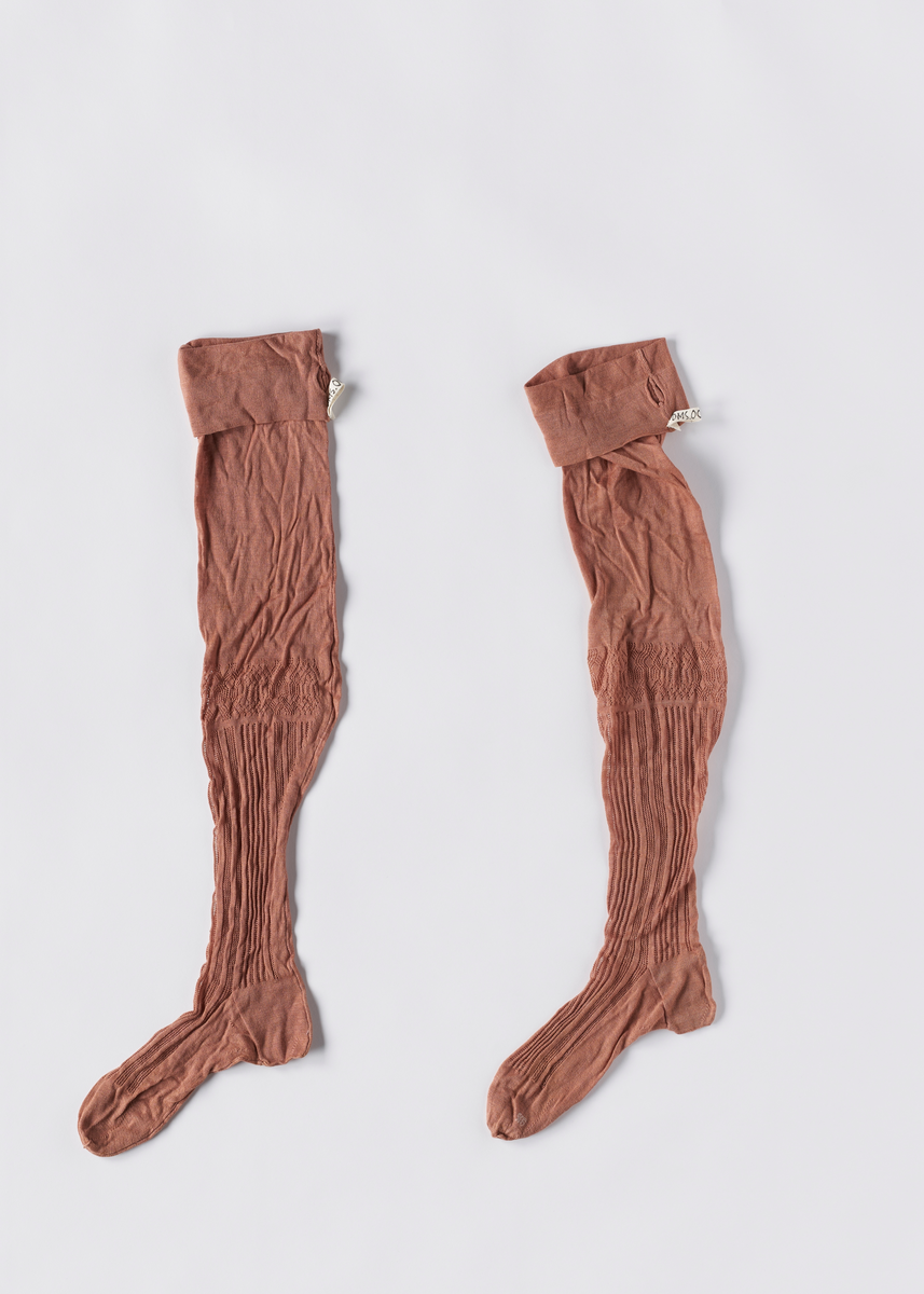 Strikkede rosa strømper i bomull. Foten er formsydd. Det er strikket hullmønster fra oversiden av foten og oppover leggen. Det er hull i brettekanten for å tre inn bånd.