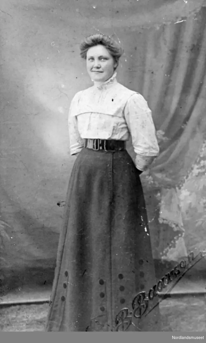 Portrett av Berntine Andersen fra Nordnesøy. Hun har hvit høyhalset bluse, mørkt sidt skjørt med belte i midjen.