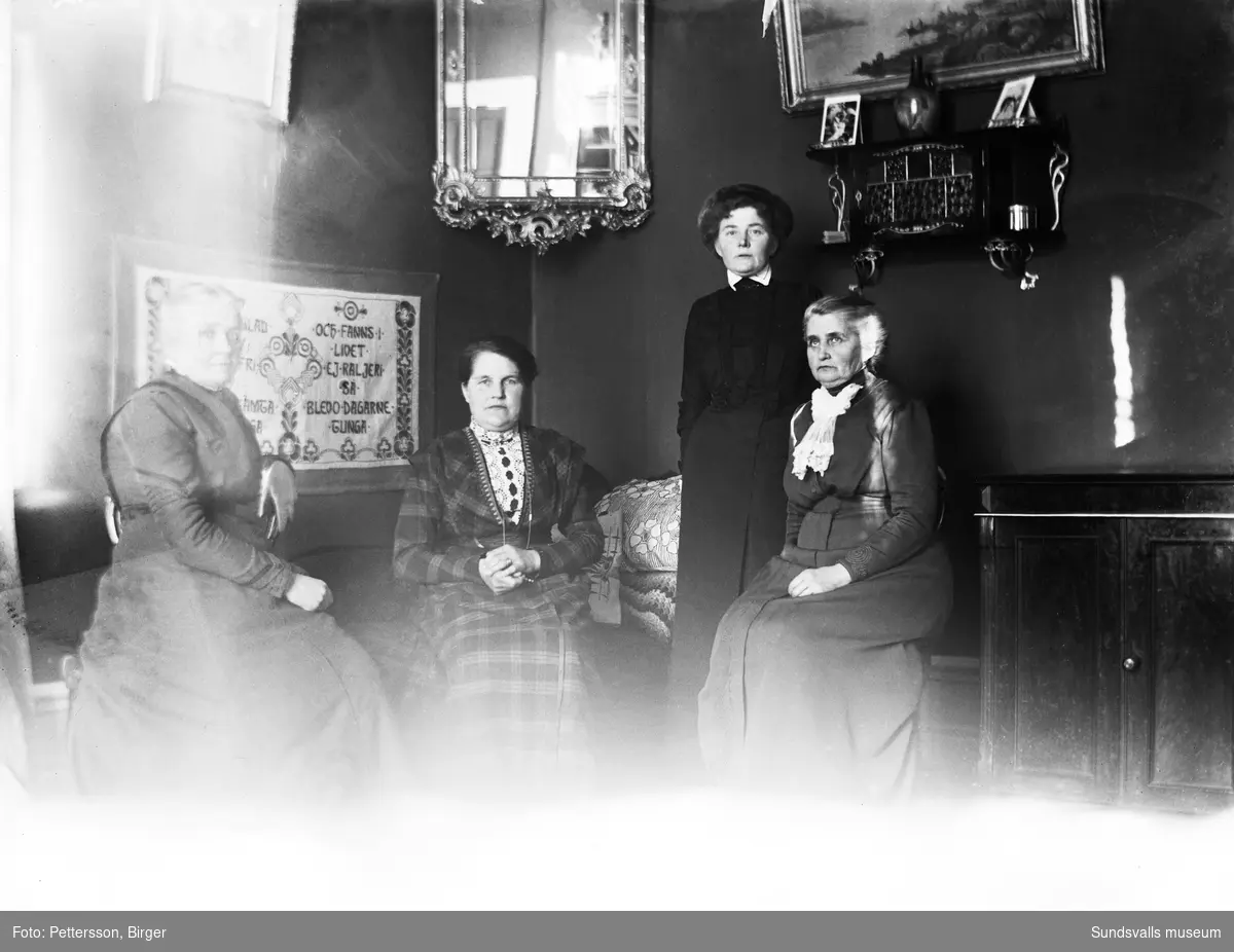 Gruppbild av fyra kvinnor som sitter i en kanapé. På väggen bakom hänger en bonad med broderade visdomsord. Kvinnan i rutig klänning är med på en bild som togs vid samma tillfälle och har nummer SuM-foto035270.