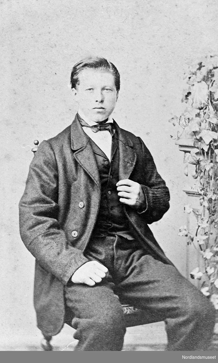 Portrett av en ung mann sittende på en stol. En blomsterdekorasjon i bakgrunnen til høyre.