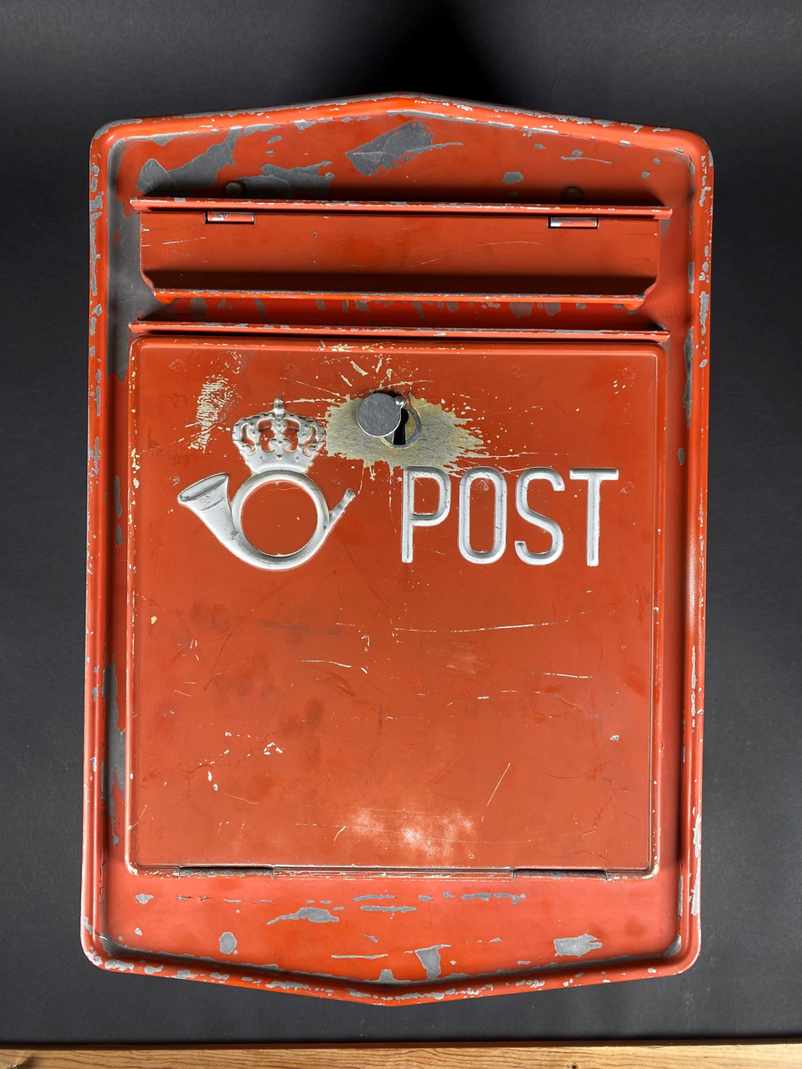 Rød postkasse med innstikksluke, merket med Postens logo og "POST". Postkassen har slitasjemerker etter bruk og noe malingsflass. Anheng for montering på baksiden.