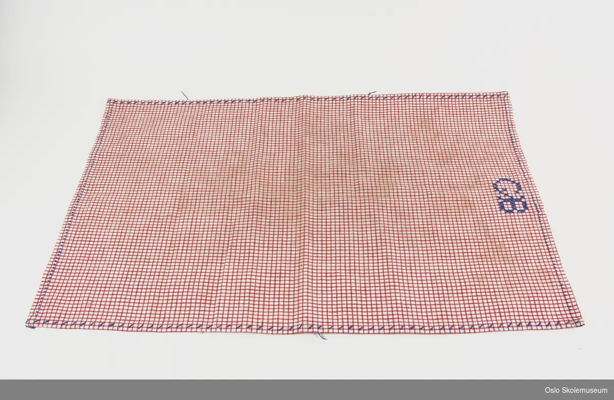 Kopphåndkle i rød- og hvitrutete tekstil. Øverst på den ene kortsiden er det brodert "GB". På baksiden er det en hempe.
