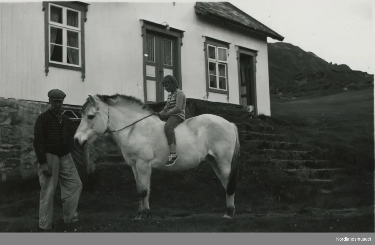 Eldre mann med en hest. Oppå hesten sitter en jente. Magnus Alfred Kristensen (1892-1969) står utenfor huset sitt sammen med hesten sin Truls. På hesteryggen sitter Magnus sitt barnebarn Marta Kristine Haug (Kristensen), f. 1951.