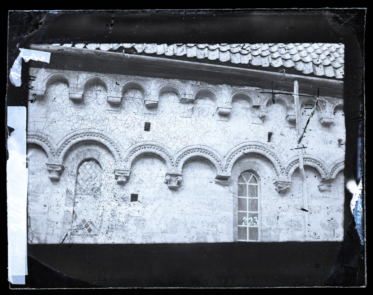 Buefrisgesims med konsoller i romansk stil. Motivet er enten nordre eller søndre tverrskip i Nidarosdomen. Bildet er tatt før restaurering, det er mange skader på konsollene, og det mangler marmorsøyler under kapitelene i første buerekke. Gamle blyglassvindu. Tverrskipet og hovedtårnet ble i all hovedsak restaurert i perioden 1891-1901.